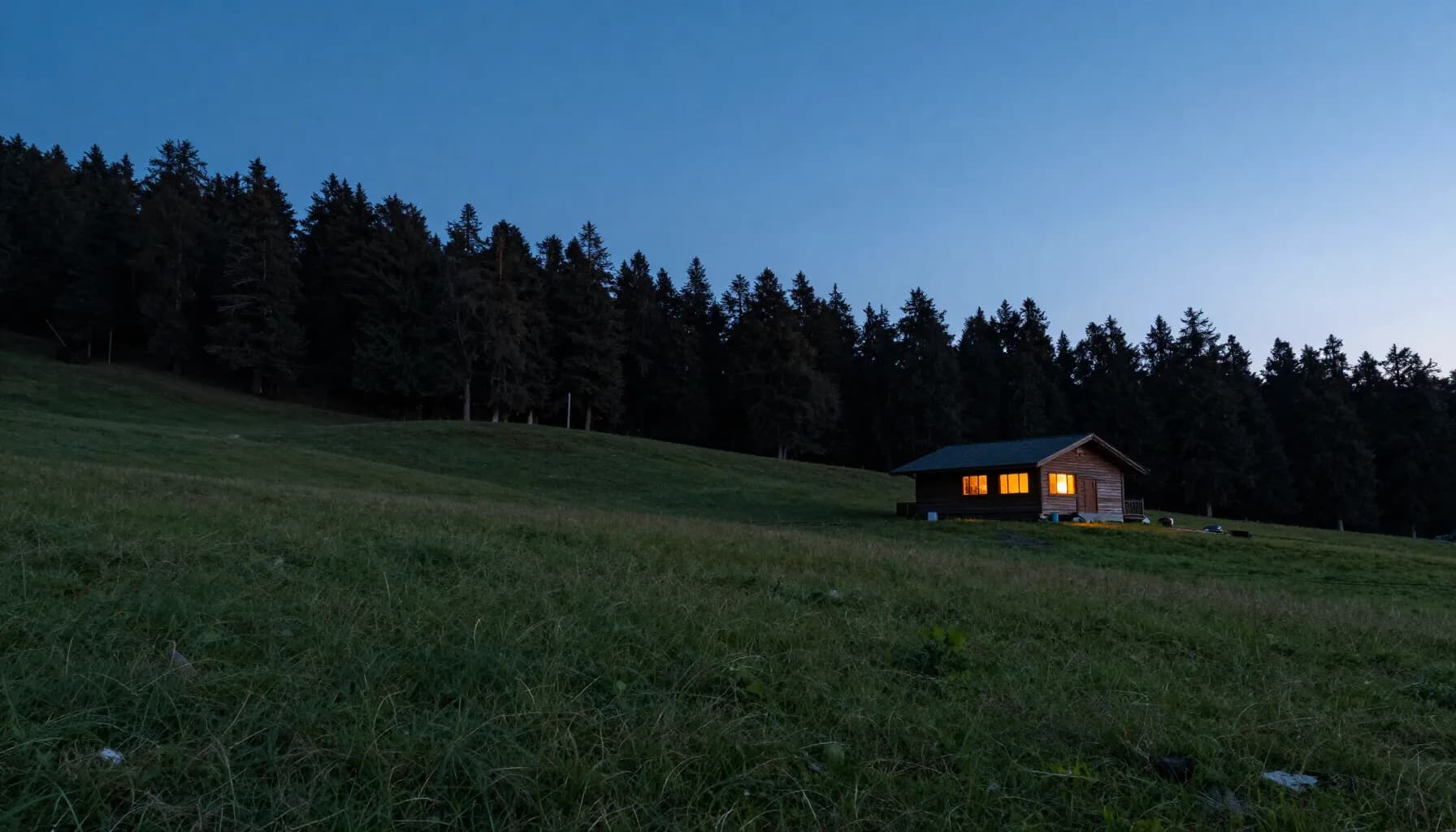 A timber lodge with lit amber windows on a dark meadow slope at dusk, with a pine ridge behind it