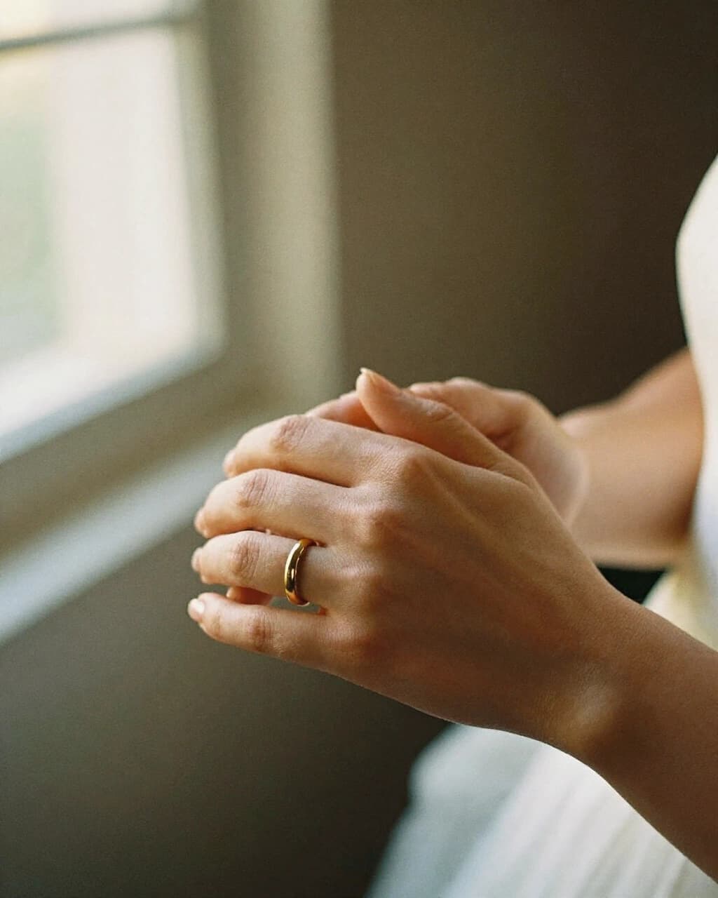 A bride's hands folded together, a gold ring on one finger, in soft window light