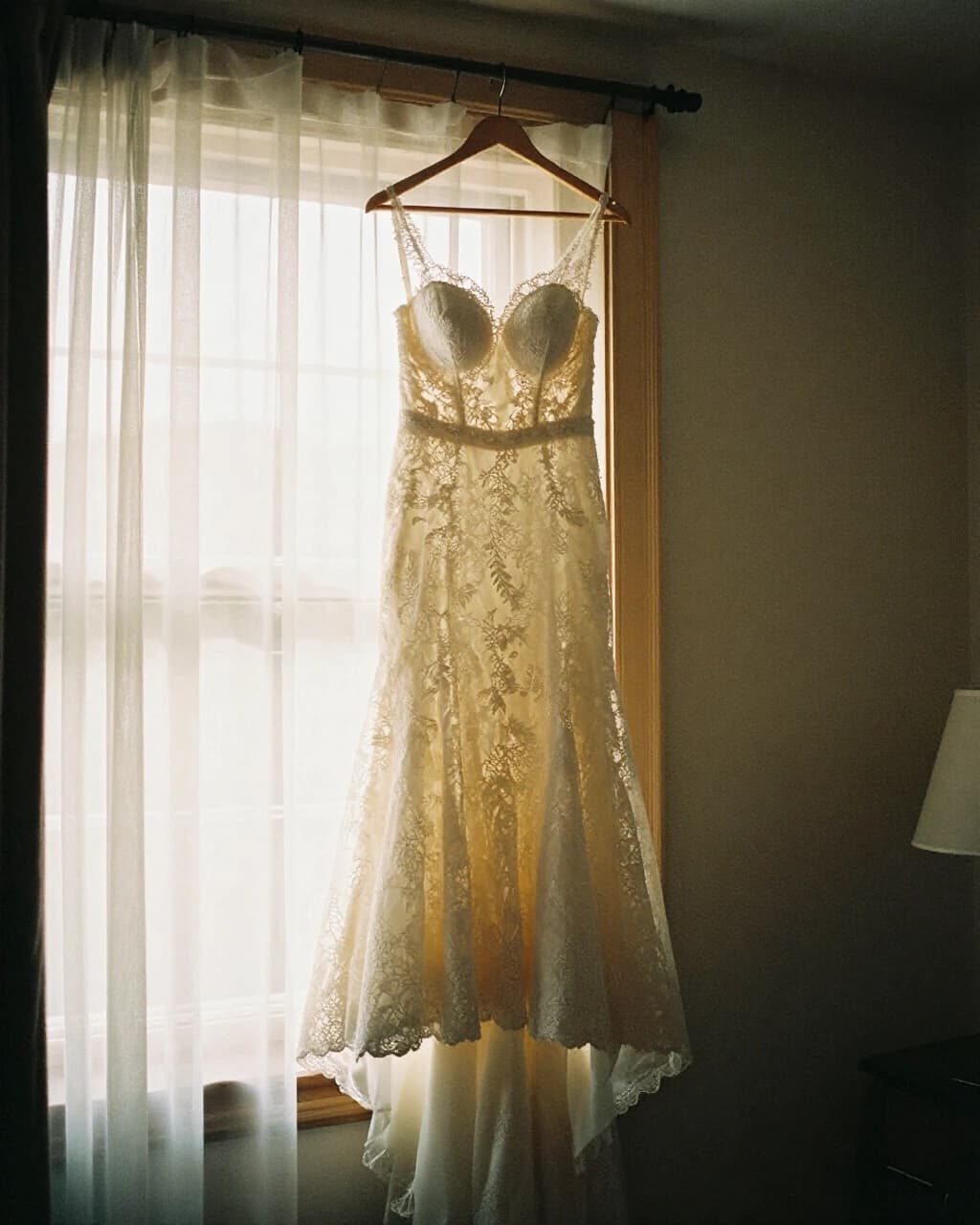 A lace wedding dress hanging in a window behind a sheer curtain
