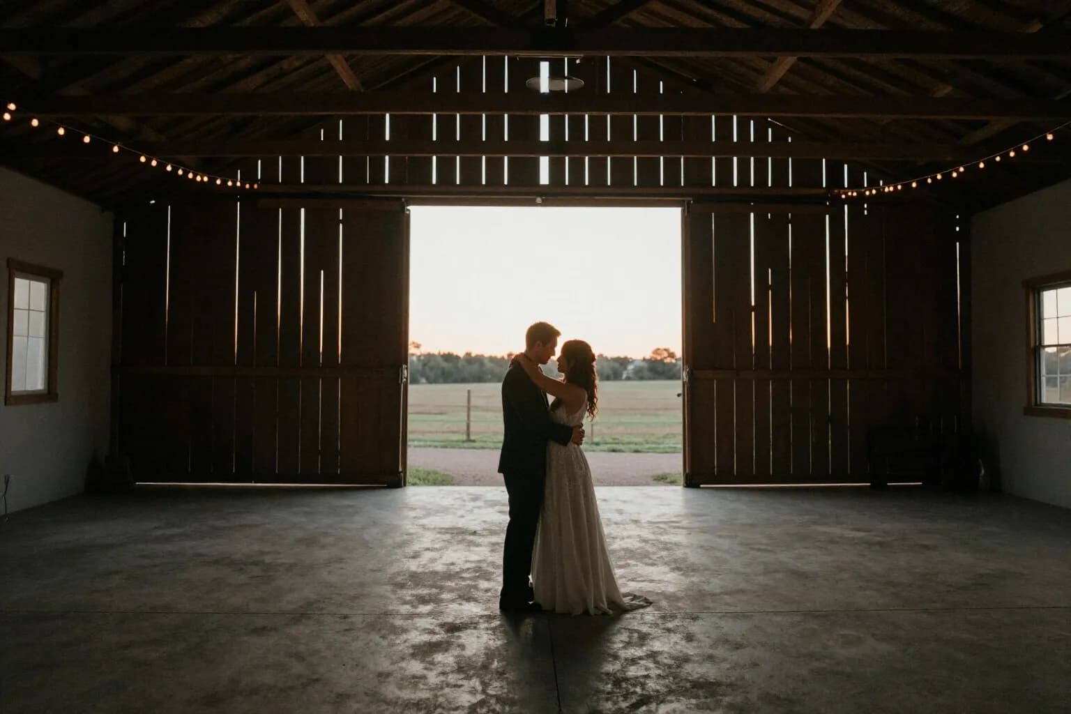 A couple dancing in silhouette in front of an open barn door at sunset, string lights above