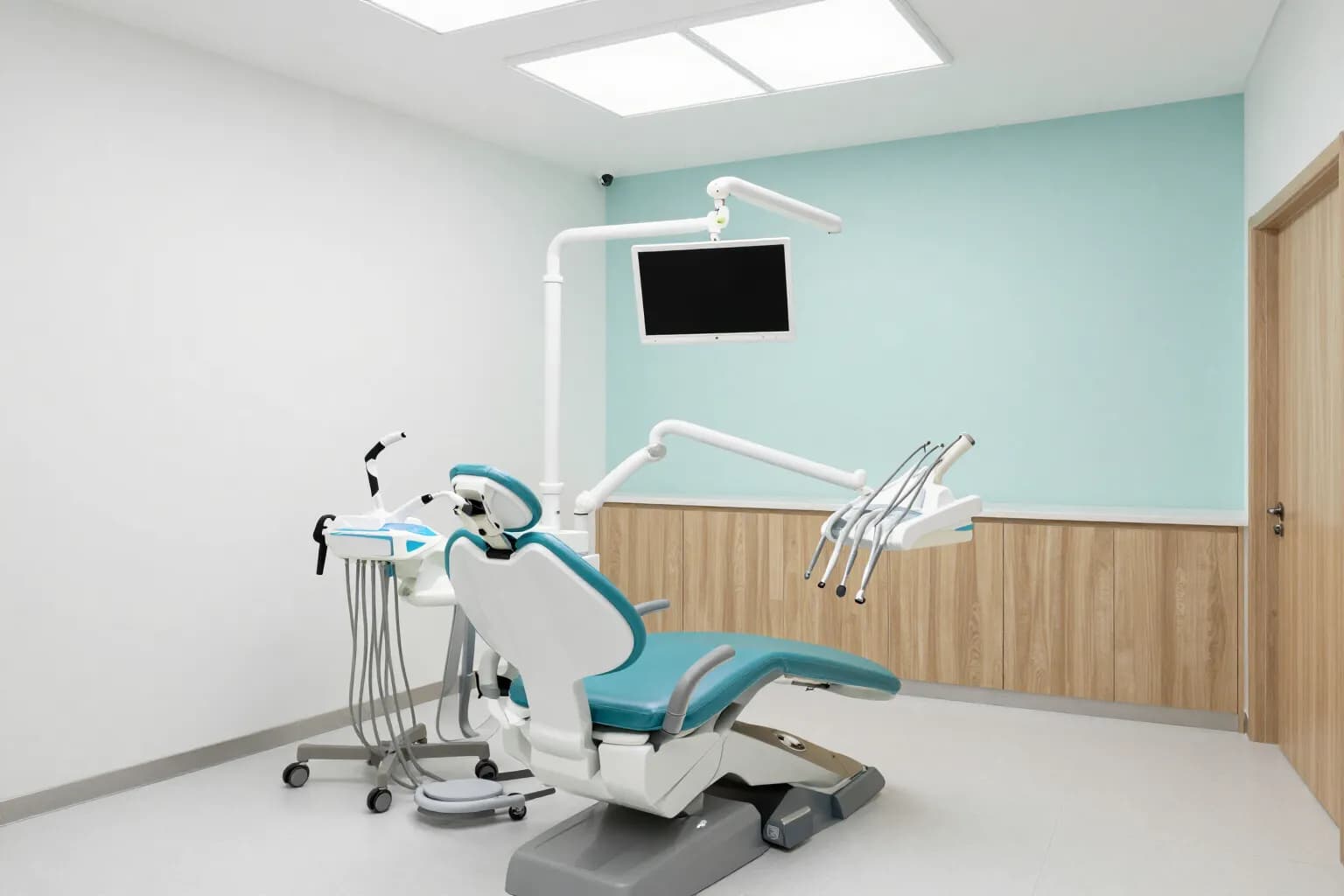 Treatment room with a dental chair, a ceiling-mounted monitor and an instrument cart
